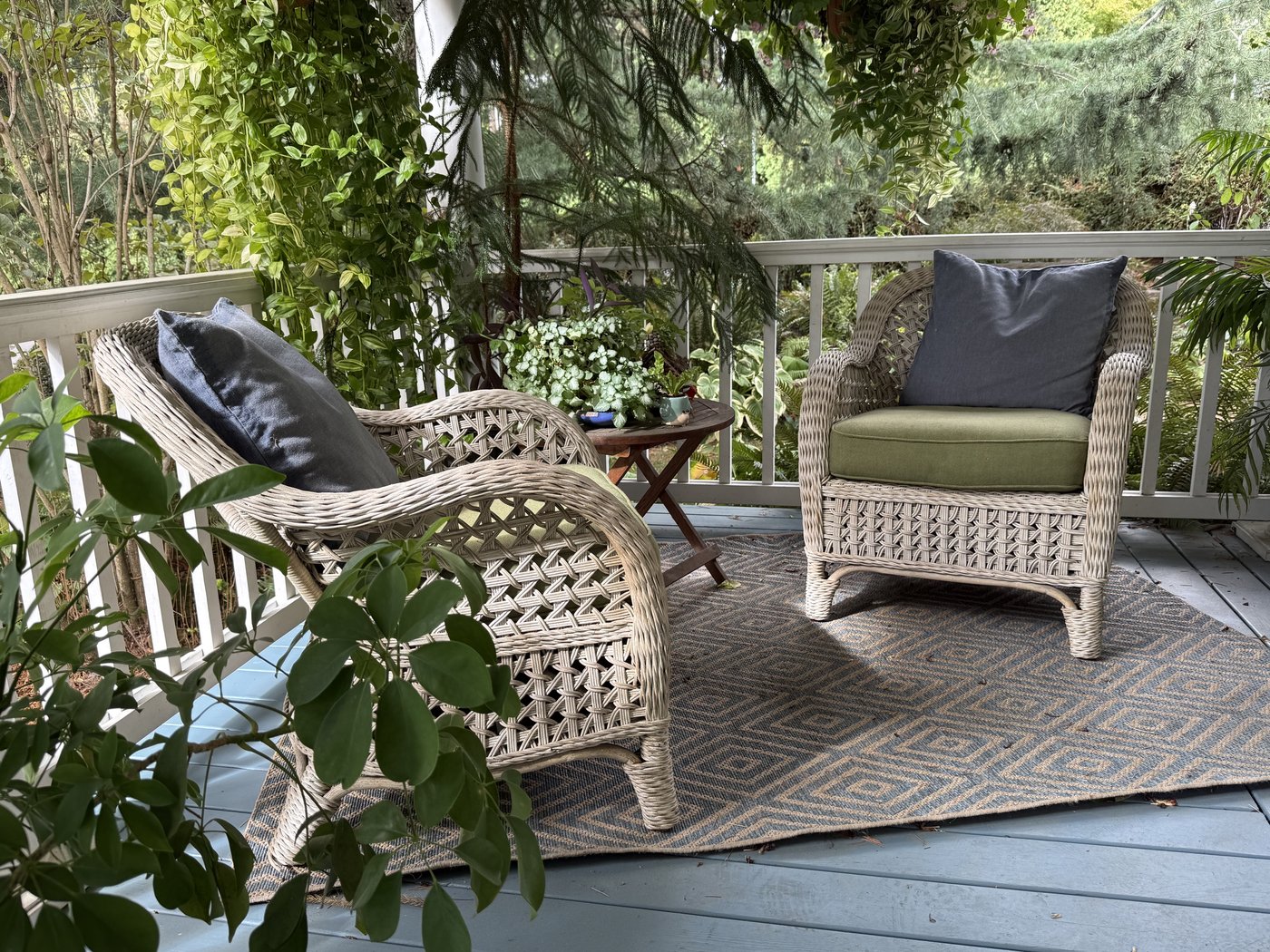 Two wicker chairs with cushions on a covered porch, surrounded by greenery