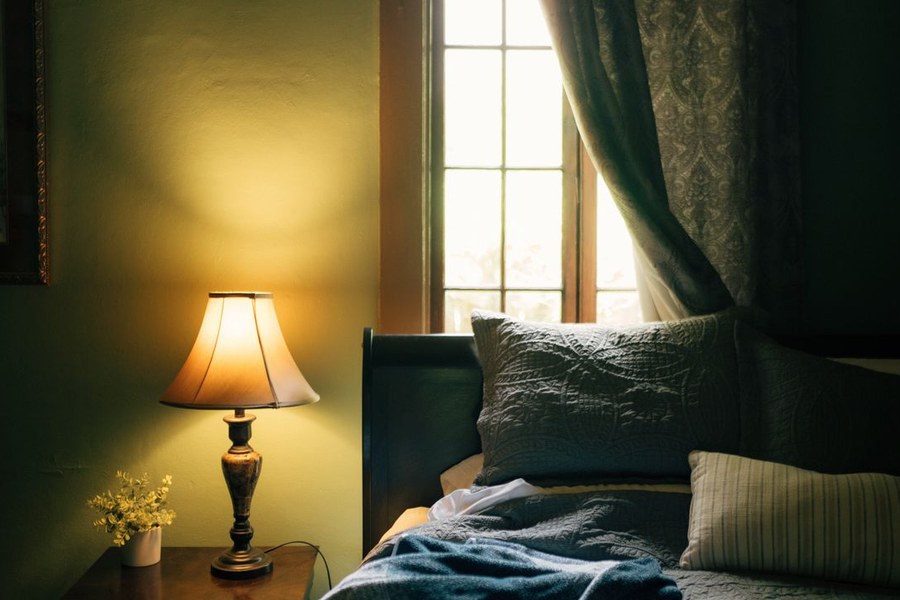 A lit bedside lamp beside a bed near a curtained window at night