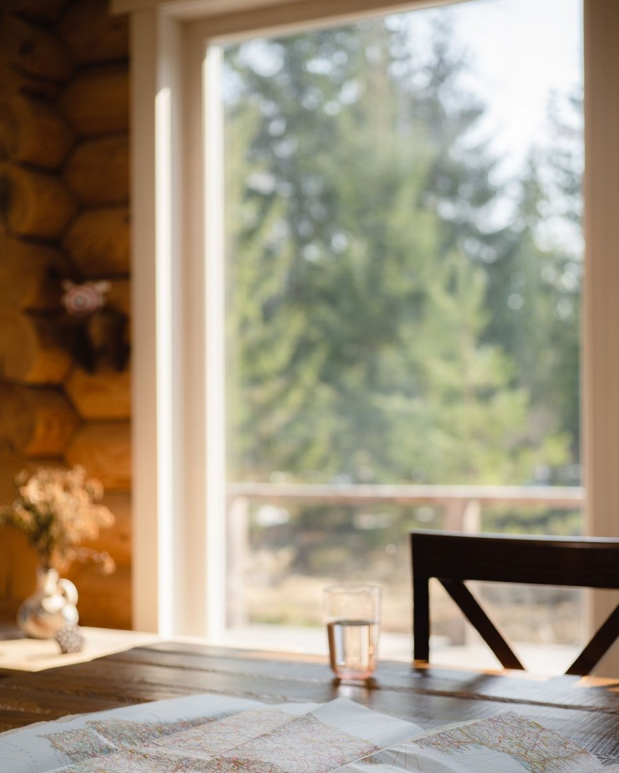 A window looking out over pine trees from inside the log home, with maps spread across the table in the foreground