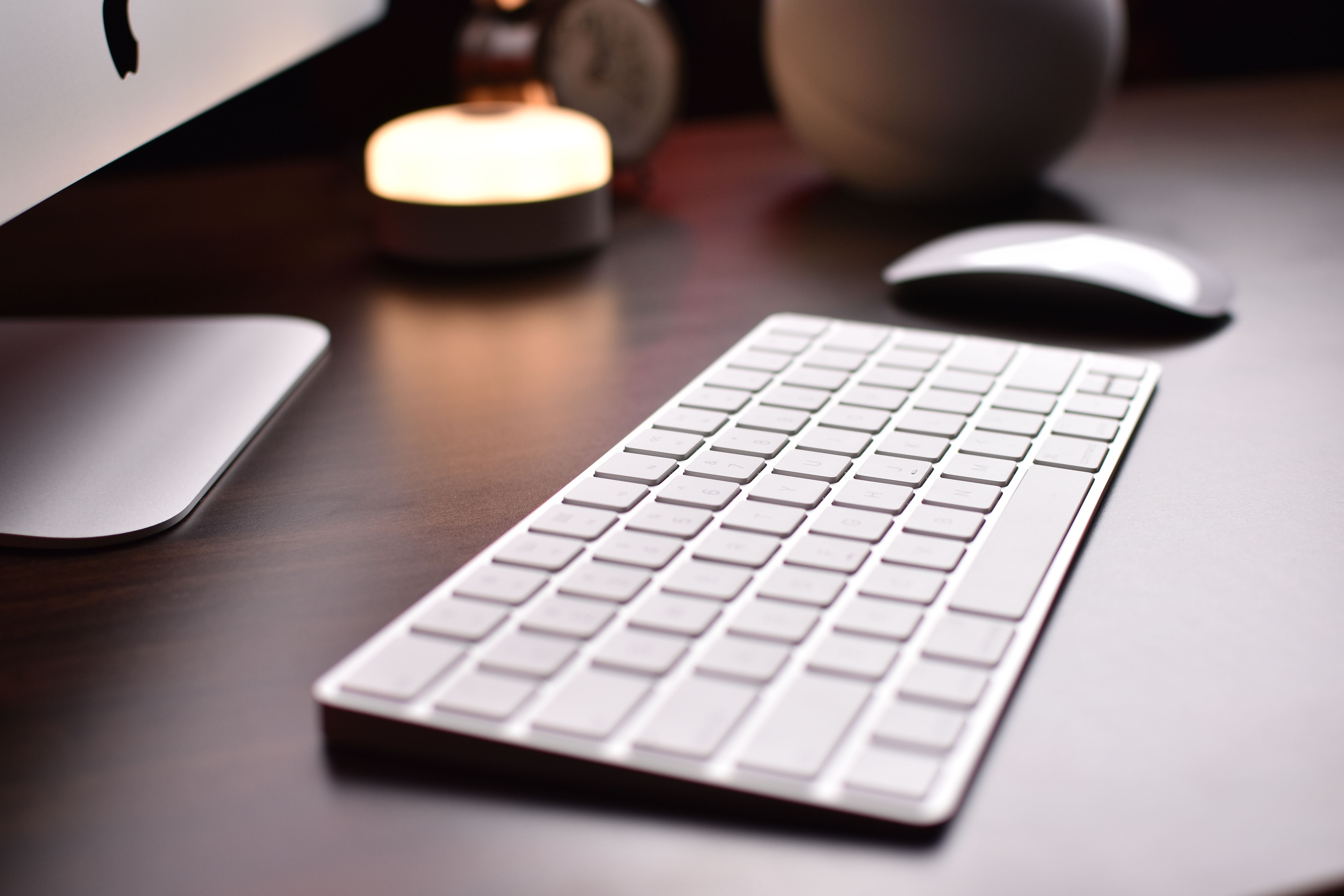A wireless keyboard and mouse on a dark wood desk, lit by a warm desk lamp at night